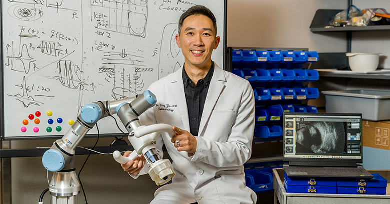 A doctor wearing a white lab coat sits holding the mechanical arm of a focused ultrasound technology. To his right, a computer shows the medical image of an eye.